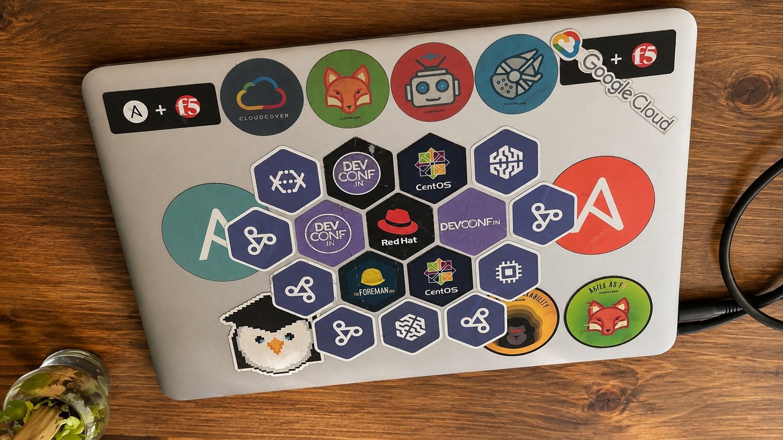 An older laptop lid tiled with hexagonal DevConf, CentOS, and Red Hat stickers plus round CloudCover stickers